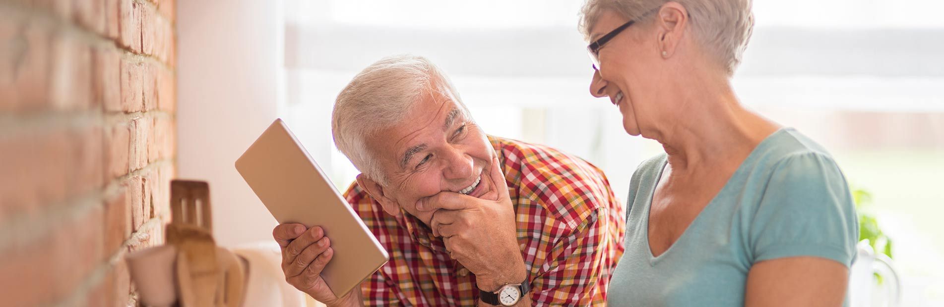 An older couple smiles at each other while one displays a tablet in a kitchen setting.